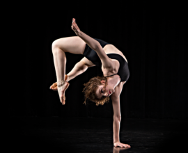 A dancer in front of a black background balances on one hand while her torso and legs curve upward.
