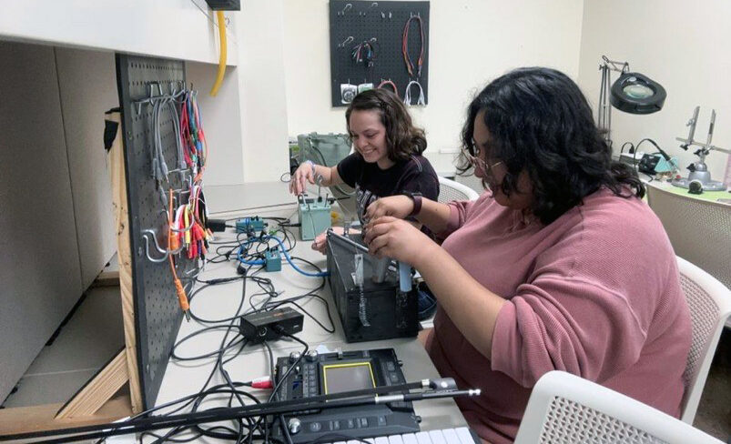 Two college students work with an intricate instrument known as a ghost box, a wooden box that has noisemaking features.
