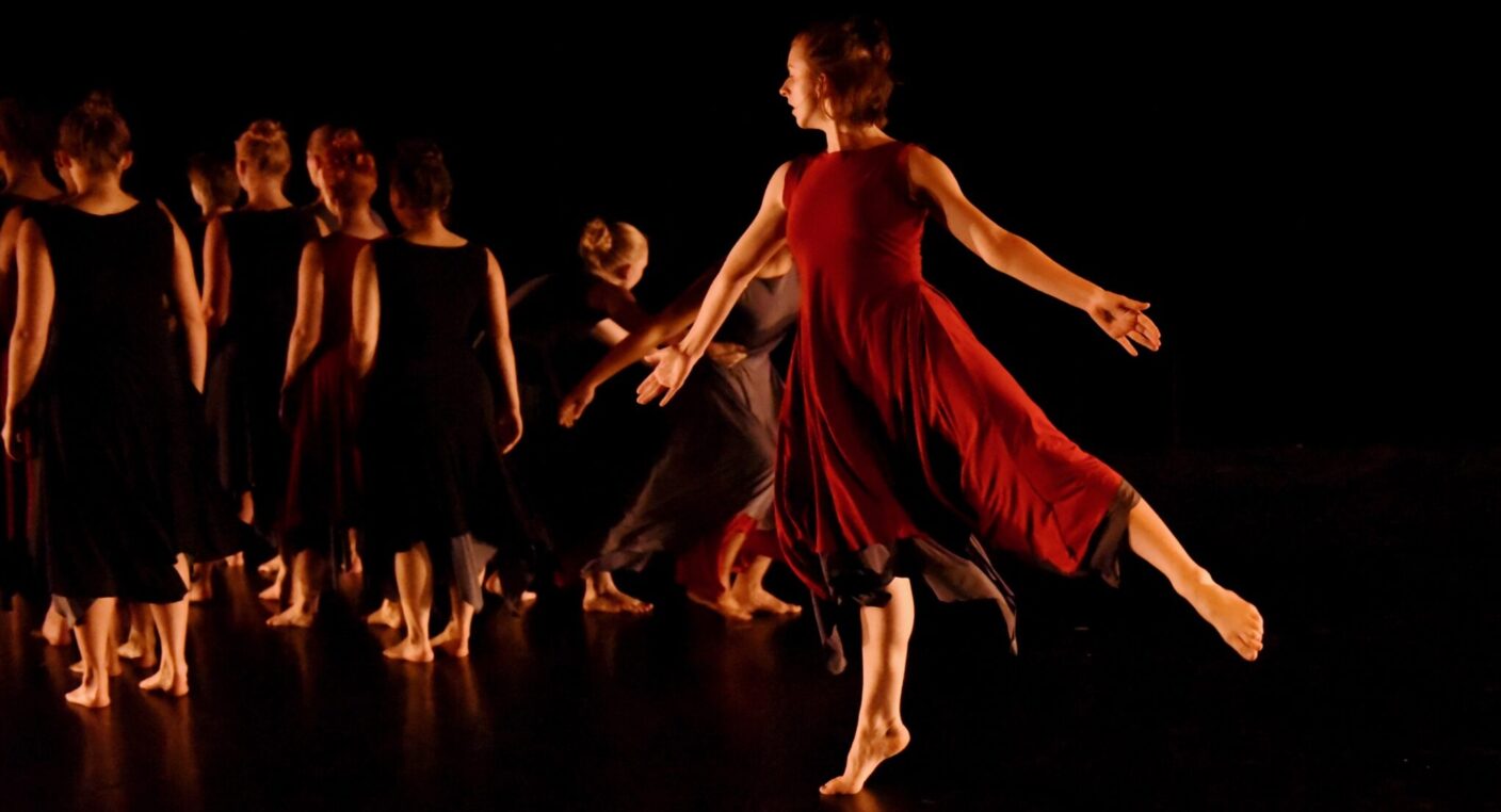 A dancer in a red dress stands on one foot and gestures with her arms as a group of other dancers in black have their backs turned.