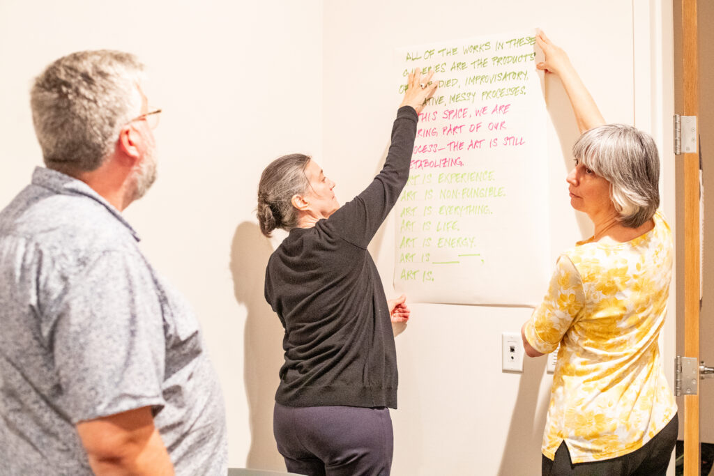 Three university professors stand in a classroom and hang up a piece of paper on a white board. 