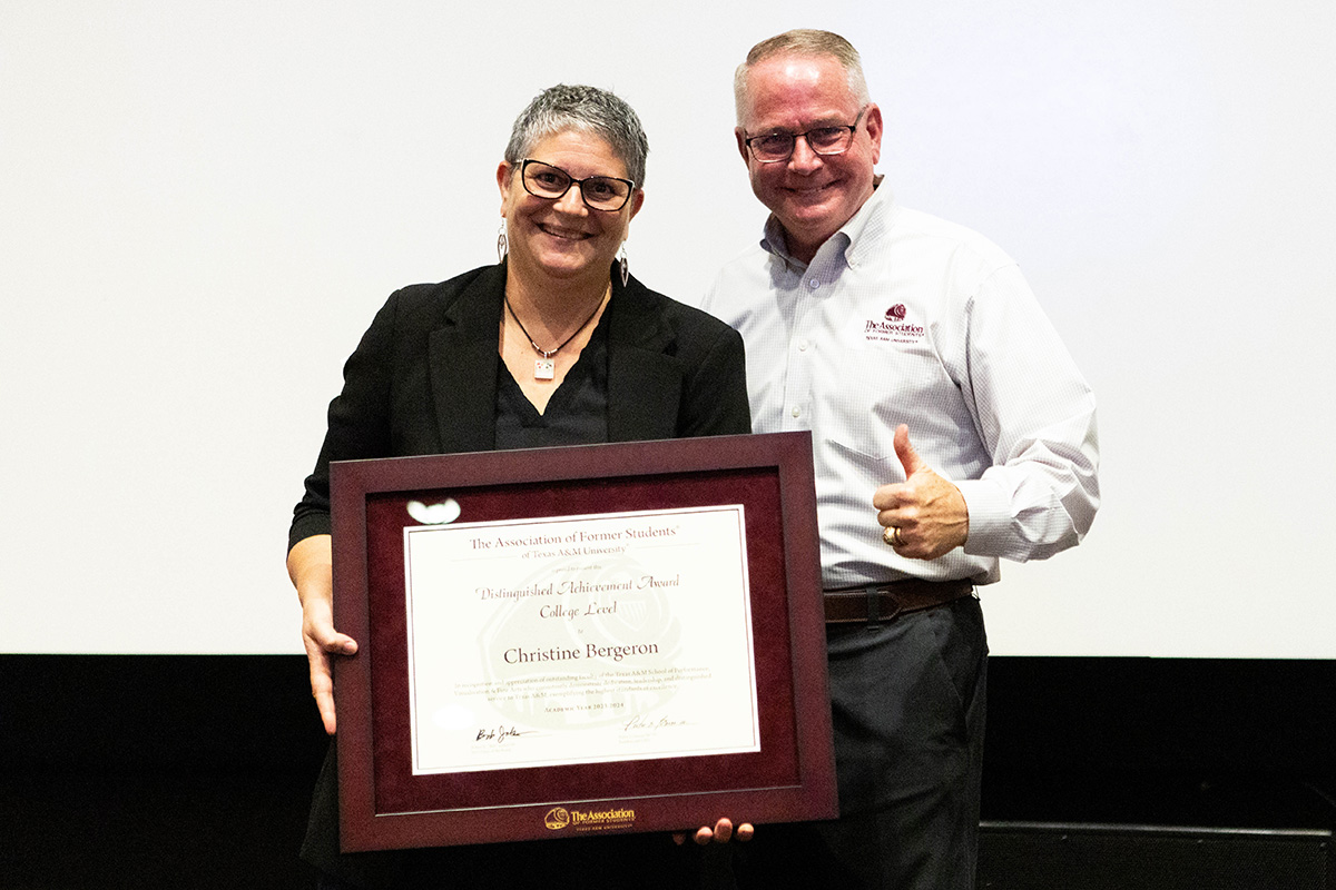 A college professor stands with a framed award while a university leader on her left gives the thumbs-up gesture.