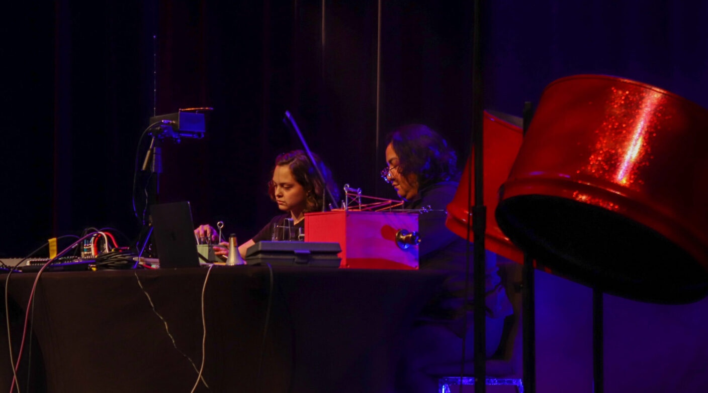 Two college students are seated at a table onstage, performing by using analog equipment.