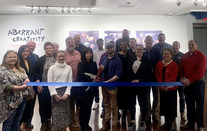 People gather for a ribbon cutting at an art exhibition, with two people holding oversized scissors by the ribbon.