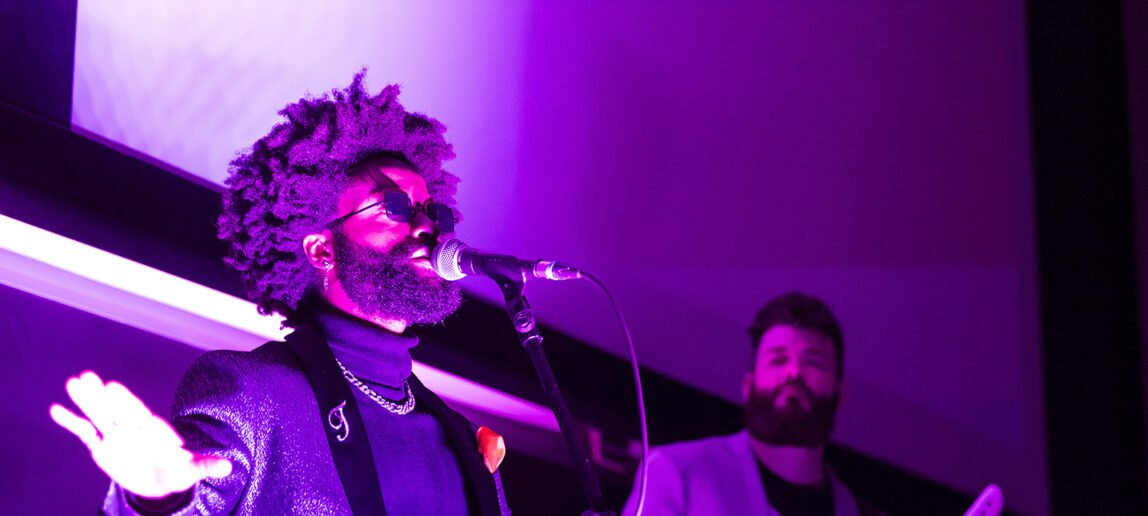 Two members of a band, one singing, one playing keyboards, perform for college students in a classroom.