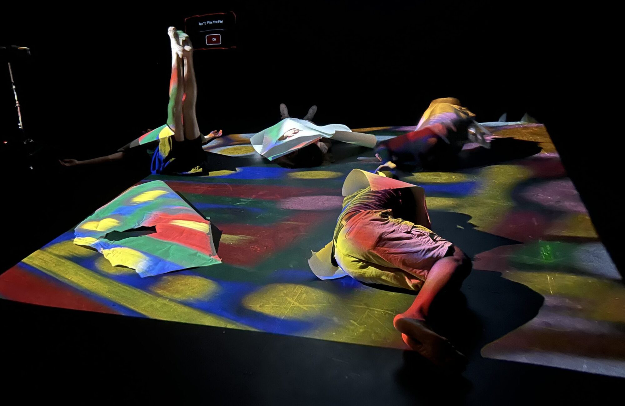 Four people lay in varying positions on the floor, which is lit in multicolored shapes from above.