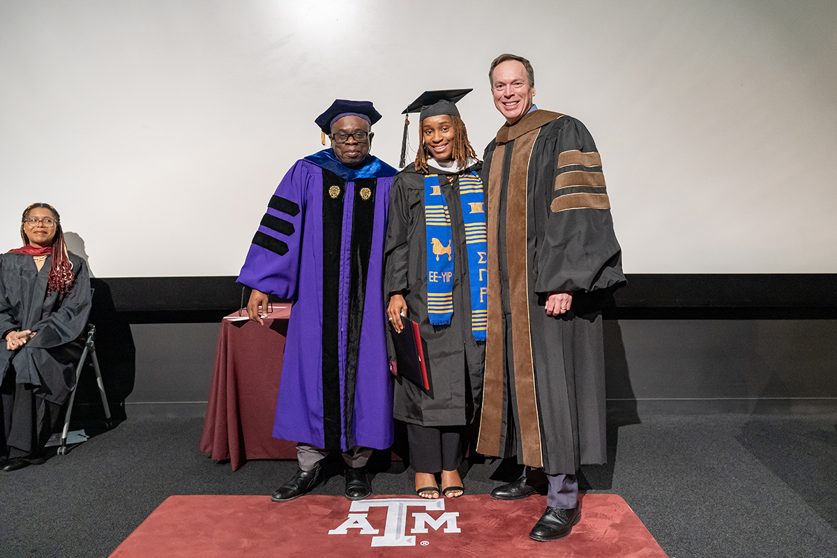 A graduating college student in a traditional cap and gown and a blue and gold sash holds a plaque on a stage. On her left is a university dean, and on her right is a university professor, all wearing graduation gowns.