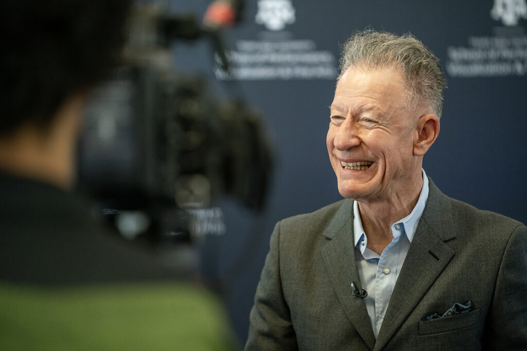 Singer-songwriter Lyle Lovett speaks to local media, in front of a backdrop that has the Texas A&M College of Performance, Visualization and Fine Arts logo.