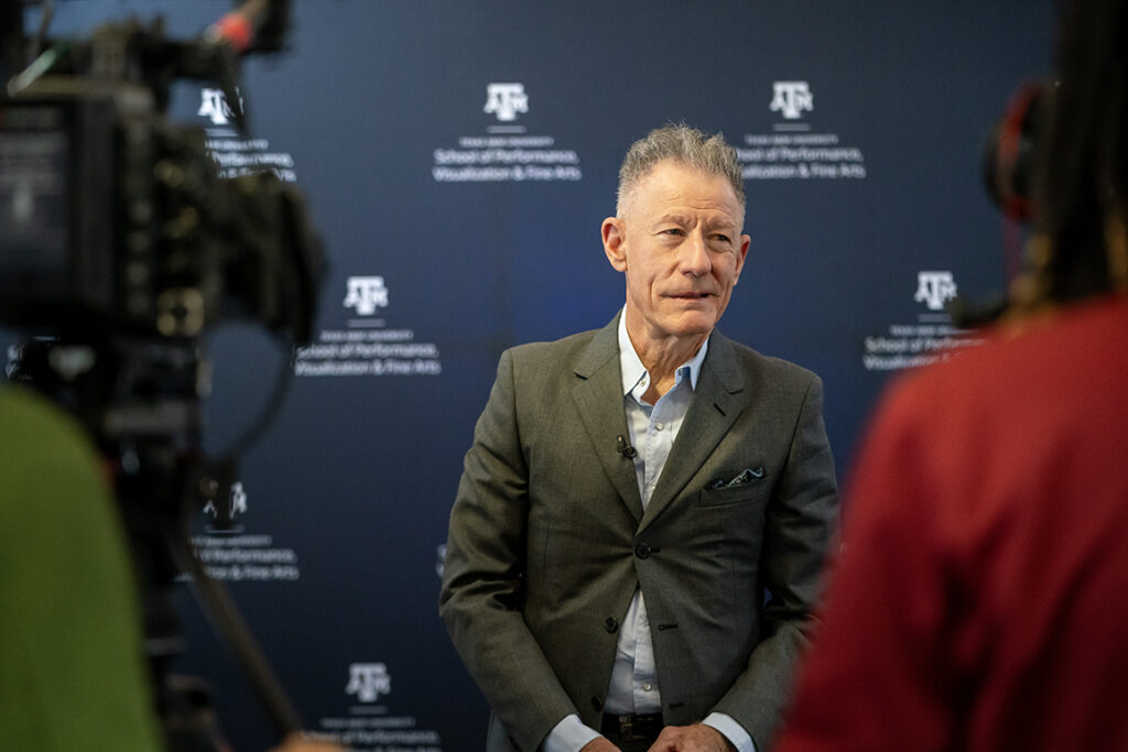 Singer-songwriter Lyle Lovett speaks to local media, in front of a backdrop that has the Texas A&M College of Performance, Visualization and Fine Arts logo.
