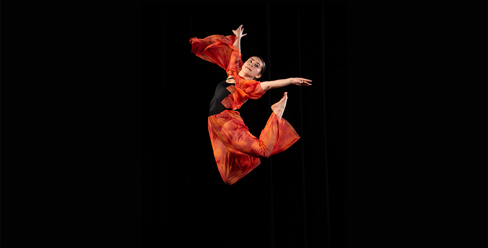A dancer wearing an orange and black outfit is seen midair, with her legs extended beyond her back, and her arms reaching outward.
