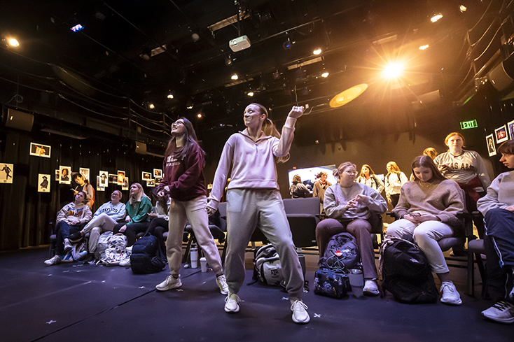 Two college dance students mirror the movements being shown on a screen, while other students are seated around them at a dance exhibit.