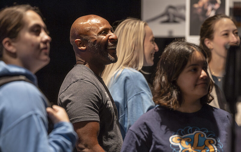 A college dance professor surrounded by students smiles as he watches a dance display at an interactive exhibit.