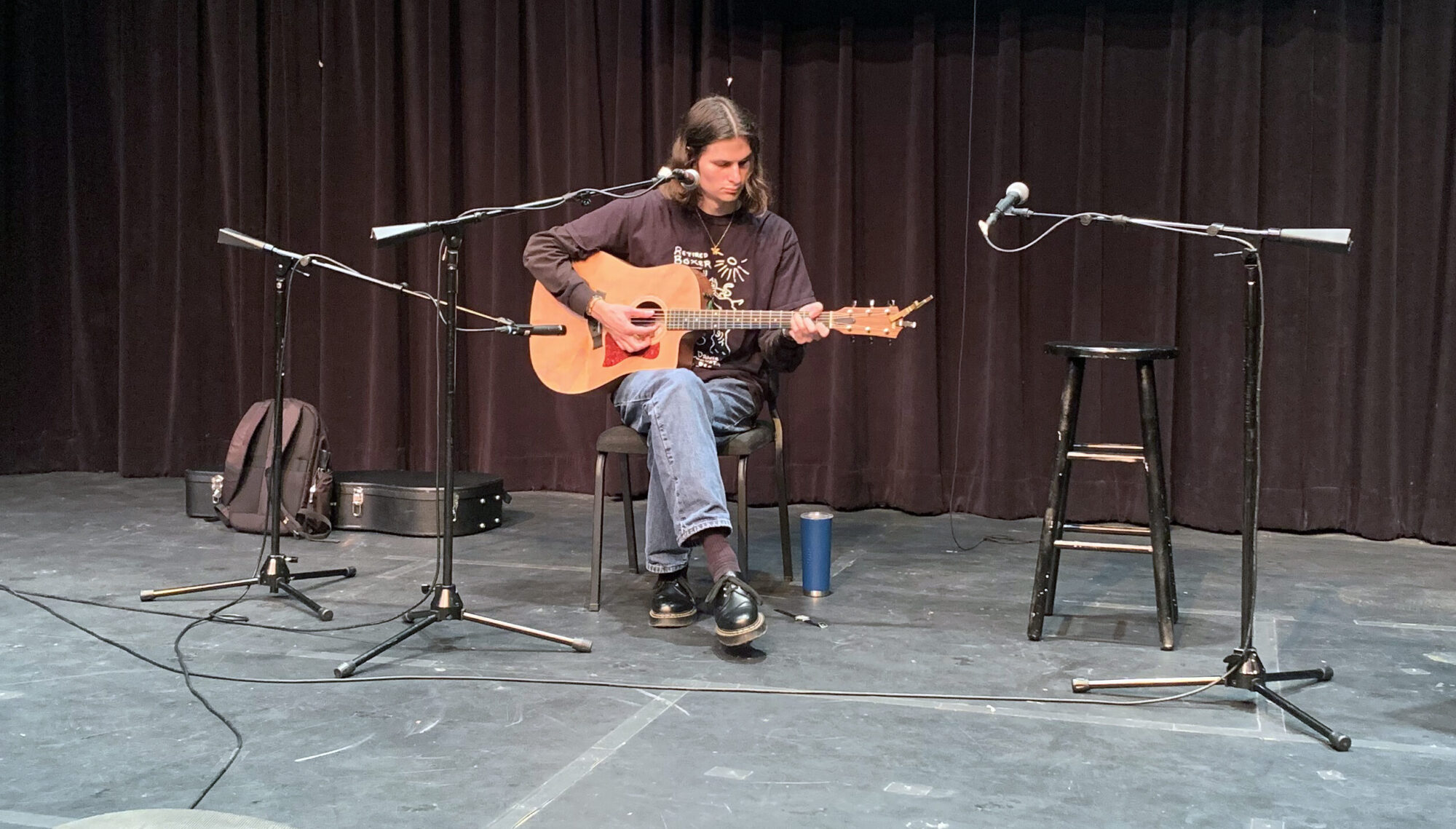 A singer-songwriter performs and plays guitar while seated on a stage.