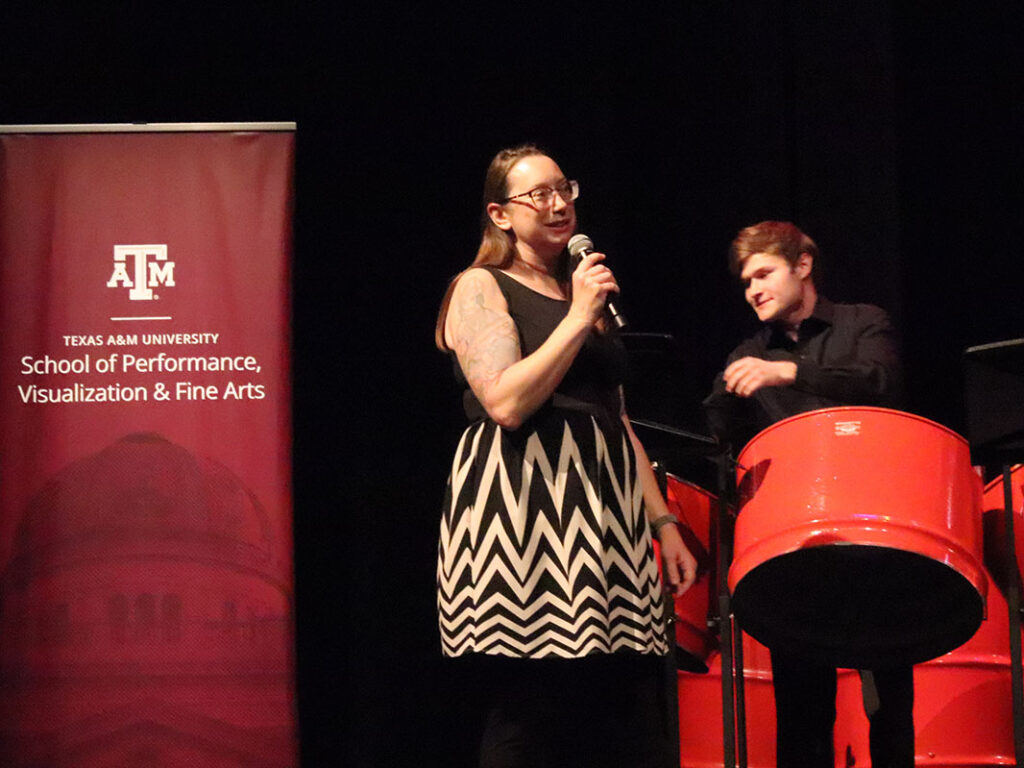 A university professor speaks into a microphone onstage. To her left is a student steel-plan player. To her right is a maroon banner that reads: Texas A&M University, College of Performance, Visualization & Fine Arts.