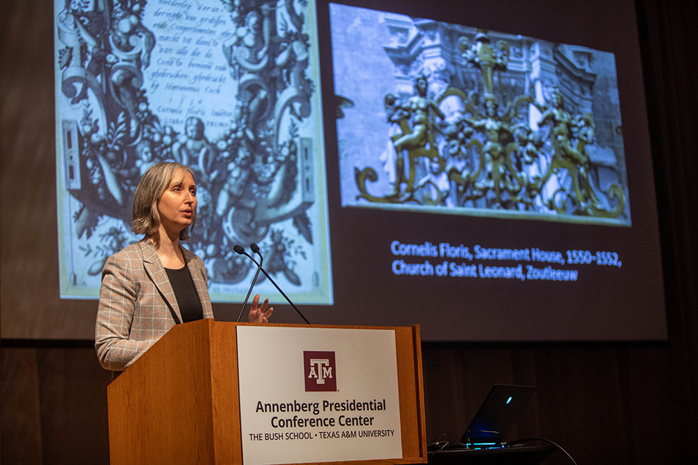 A college professor stands at a podium giving a presentation. On the screen behind her are two images of ornament title prints.