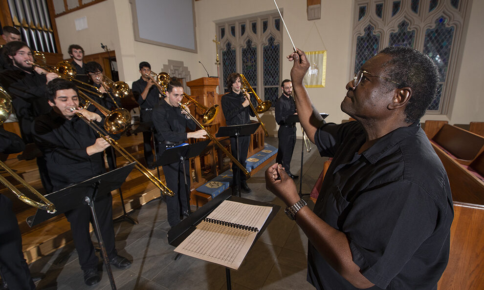A music professor standing on the right conducts a choir of trombone players.