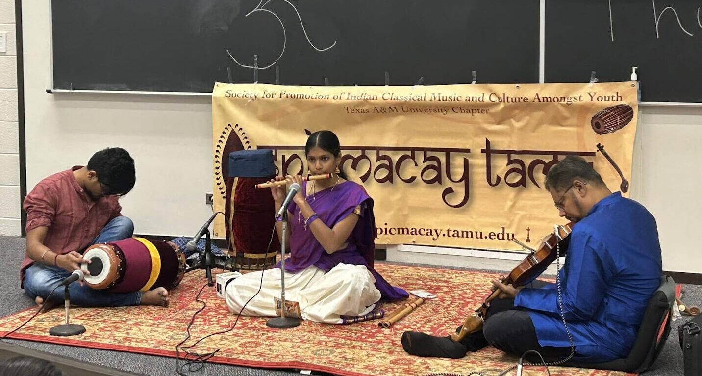 Three students play classical Indian instruments while sitting on the floor of a classroom.
