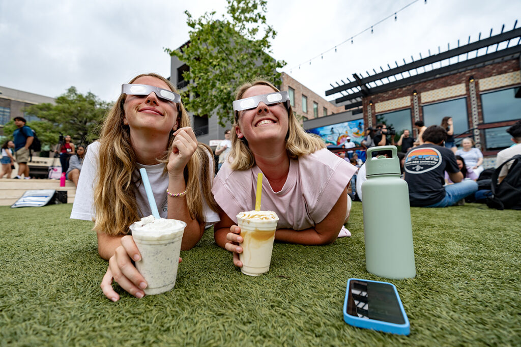 Two students laying on the grass and wearing solar glasses smile as they look toward the sky.