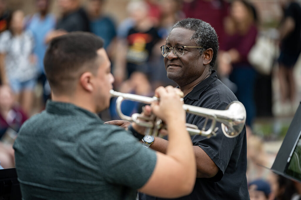 A trumpet player performing with other musicians in front of a group of people. We can also see his conductor.