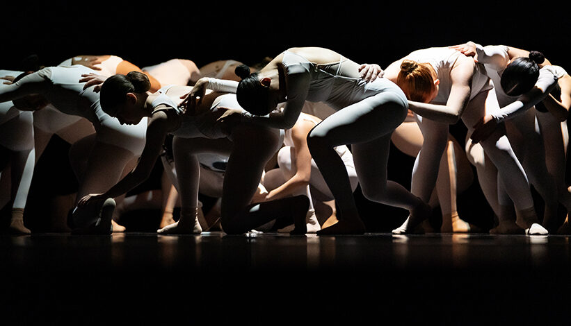 Members of a high school dance troupe, all wearing white, are shown performing onstage. They are all crouched down toward the floor, with their arms on the dancer in front of them.