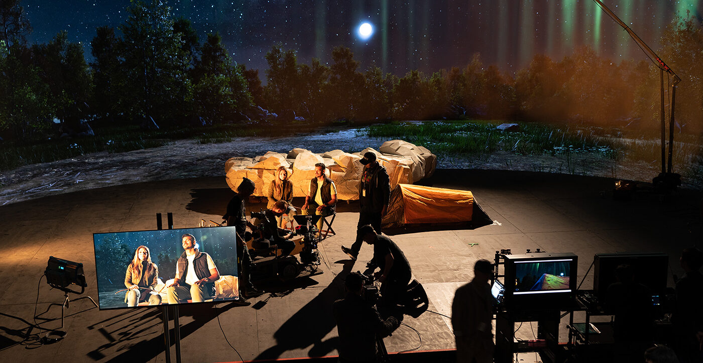 Two actors are on a virtual production stage shooting a movie. The camera view is visible on a screen to the left. The images behind them make it appear they are in a nighttime wilderness scene. Crew members are shown working in the background.