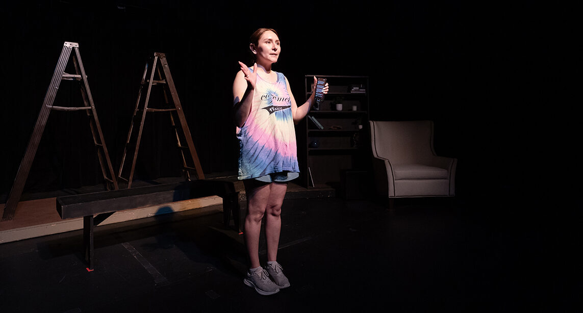 A college student who serves as the director of a musical stands on a stage as she addresses her cast and crew.