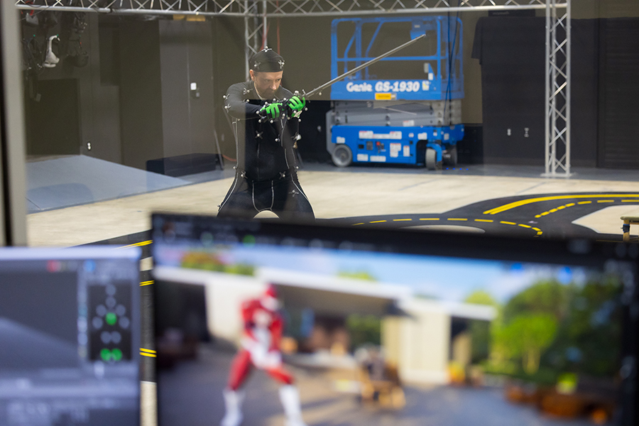 A man wears a black bodysuit outfitted with markers in a motion capture facility. He is holding a sword up with his hands. In the foreground, a computer screen shows an avatar that is mirroring his movements.