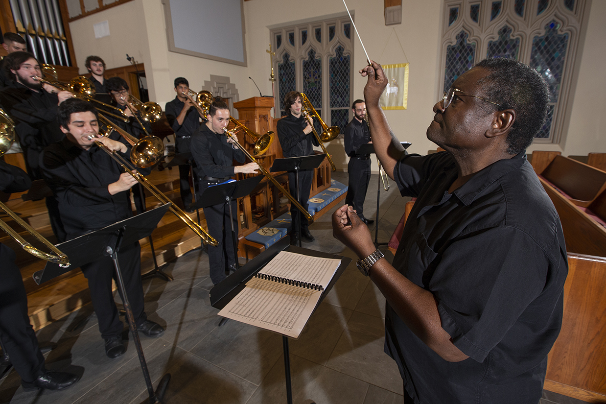 A music professor conducts an orchestra of trombone players in a church setting.