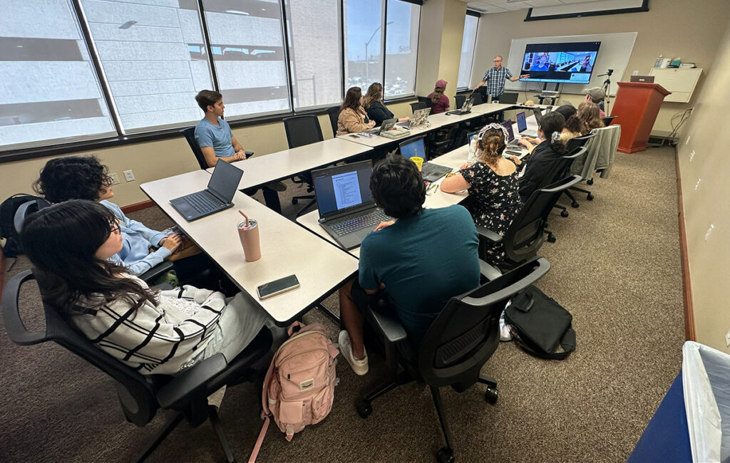 College students are seated around a long rectangular arrangement of tables next to a wall of windows. The students are listening to a lecturer speaking at the end of the table. Behind the lecturer is a screen showing another person joining the conversation via Zoom.
