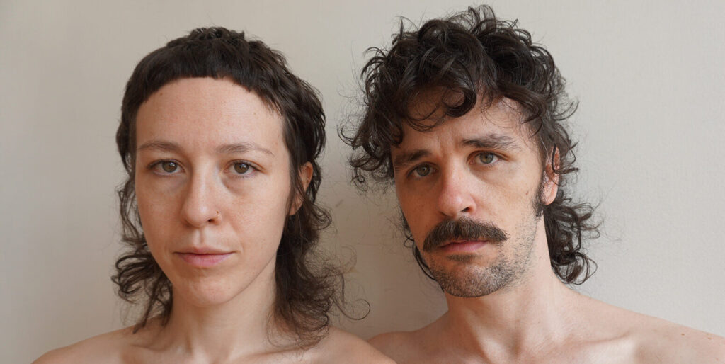 Two performance artists stand for a portrait in front of a grayish background. Both are shirtless from the upper torso.