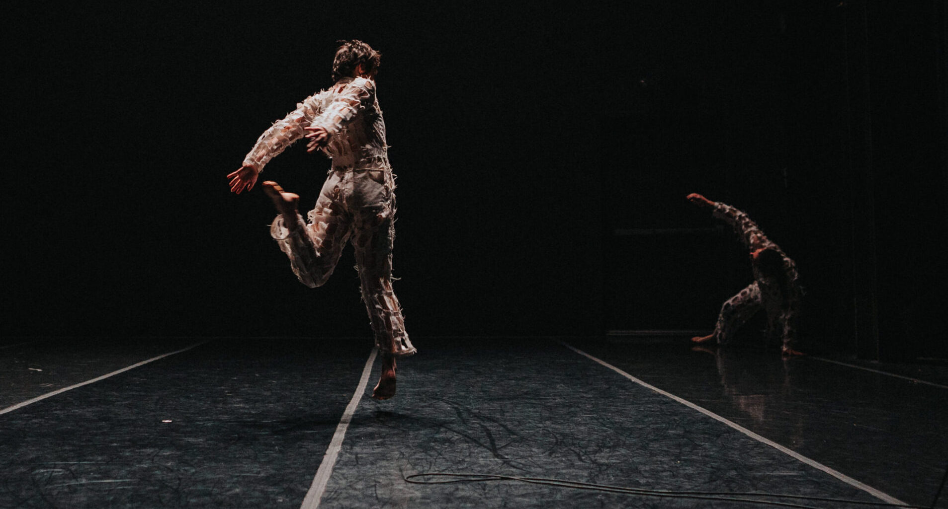 A dancer is shown from the back in a darkened stage environment. The dancer is leaping off their right foot, with their left leg bent behind them. To the right another dancer performs, balancing on their left arm and leg.