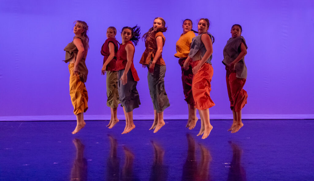 Seven dancers are seen jumping in the air in unison on a stage with a purple-lit background.