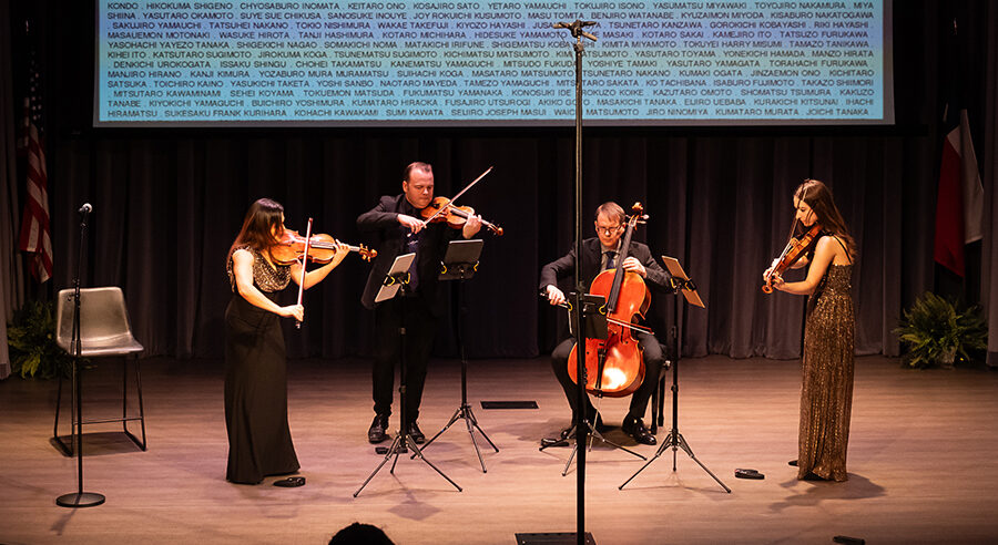 A string quartet performs onstage. Behind them, projected on the screen are the names of Japanese-Americans who were incarcerated during World War II