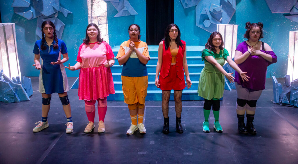 Five student actors, all dressed in a different color (green, pink, blue, purple and orange) perform on stage in a theatrical production.