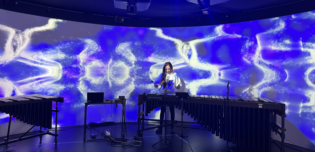 A musician plays the marimba in front of a 360-degree screen showing blue and white patterns and designs.