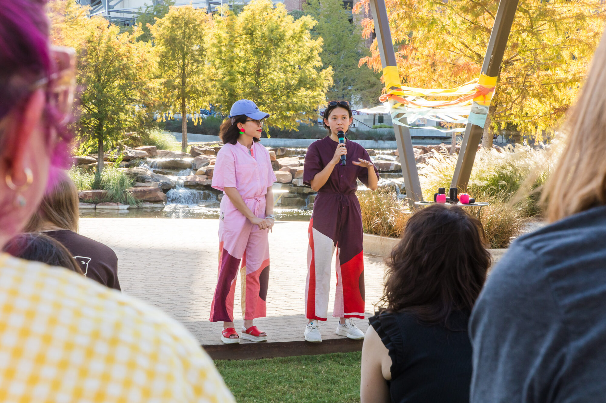 Two women stand on a stage in an outdoor setting. One is speaking into a microphone.