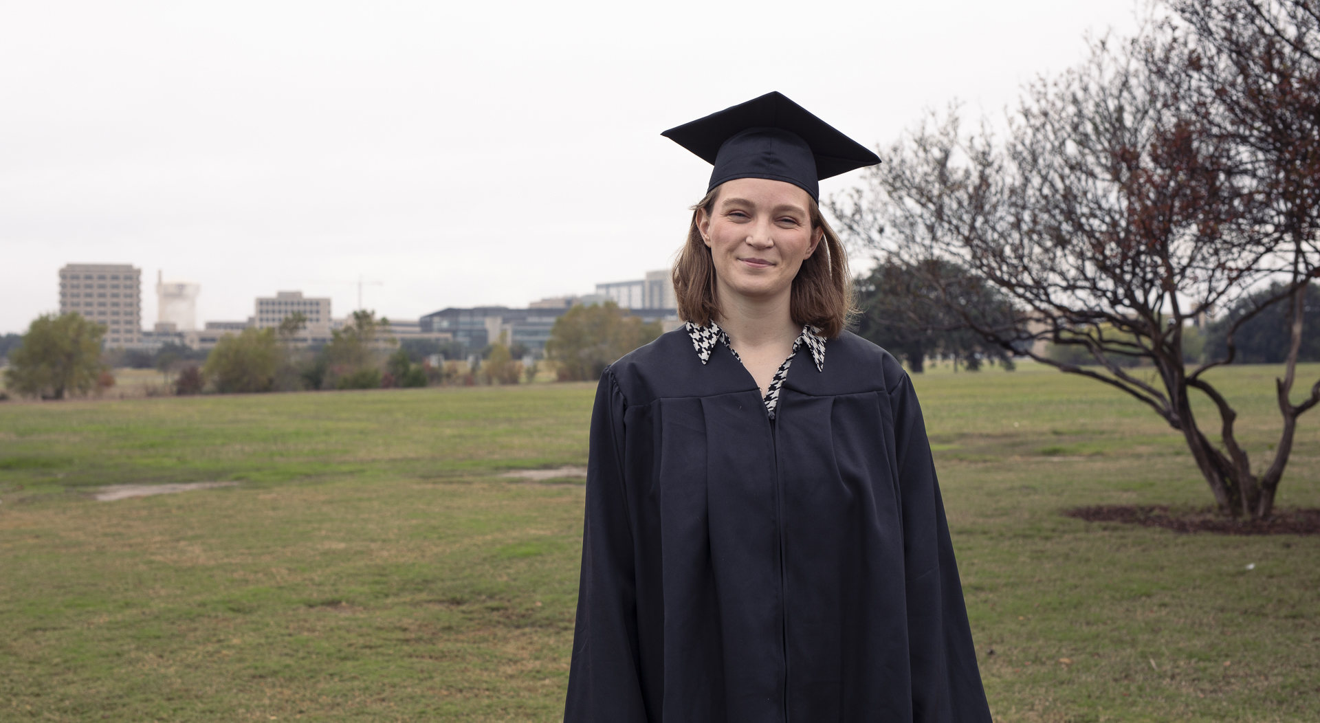 A college student stands on a campus, wearing a traditional cap and gown.