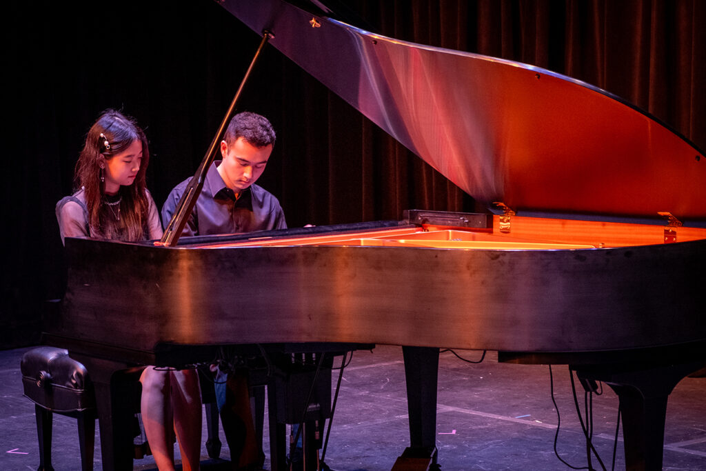Two college students sit together at a piano as they perform for an audience.