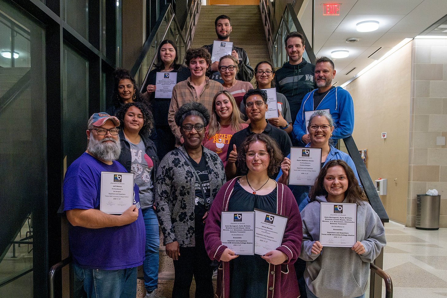 College students, faculty and staff members stand on a stairwell. Most are holding certificates of recognition.