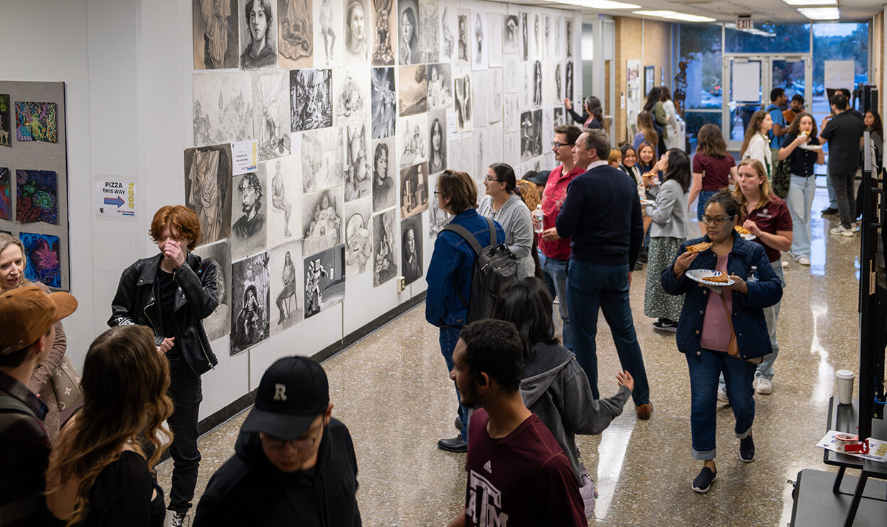 A college hallway is filled with people looking at artwork displayed on the walls.