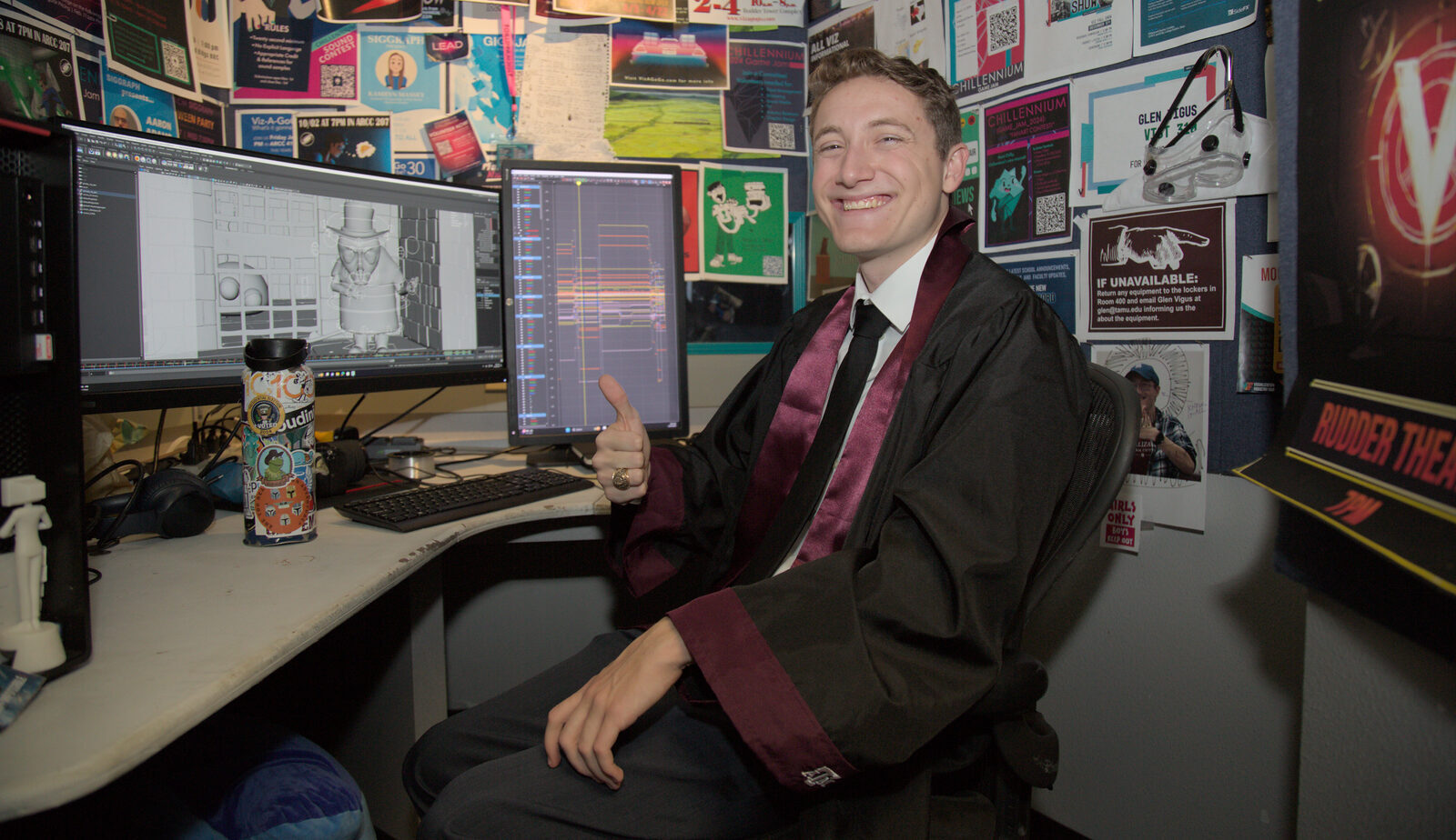 A college student smiles and gives the thumbs-up gesture while sitting at a cubicle with his computer, wearing a graduation gown.