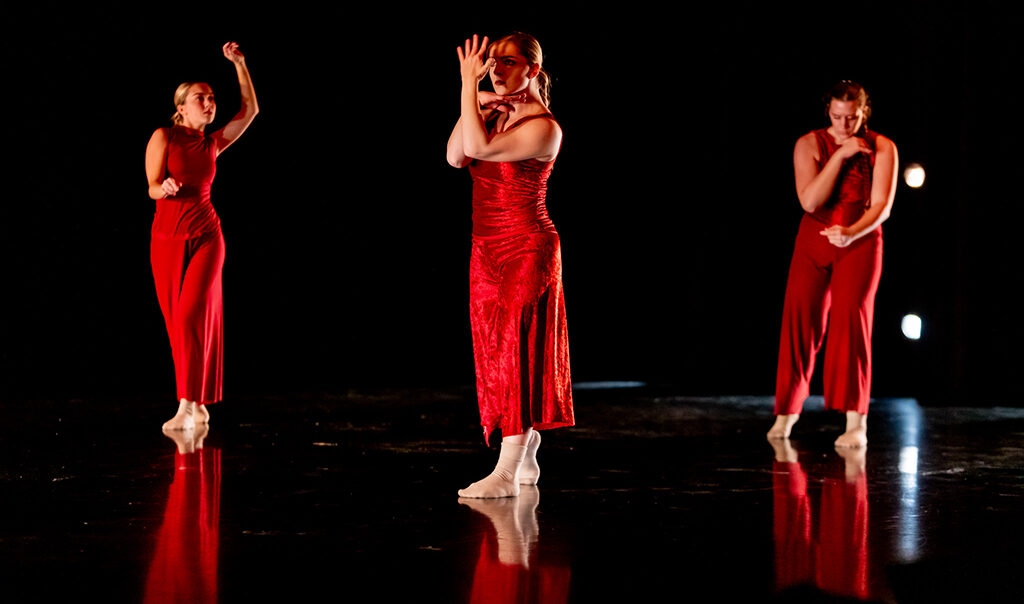 Three college students wearing red outfits perform on a darkened stage.