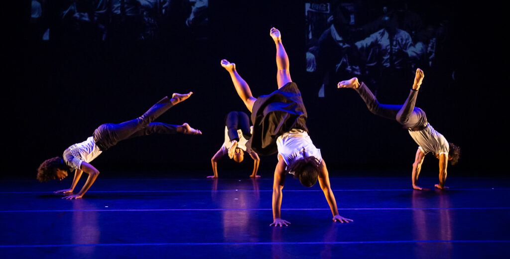 Four dancers have their hands on the stage floor and their bodies and legs extended upward.