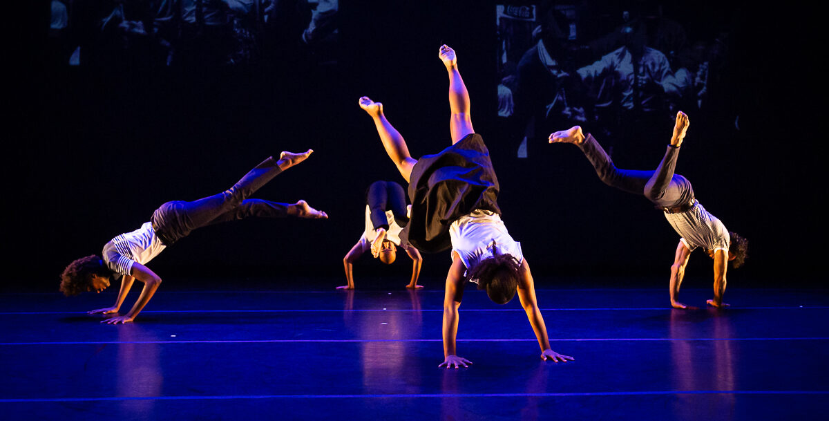 Four dancers have their hands on the stage floor and their bodies and legs extended upward.