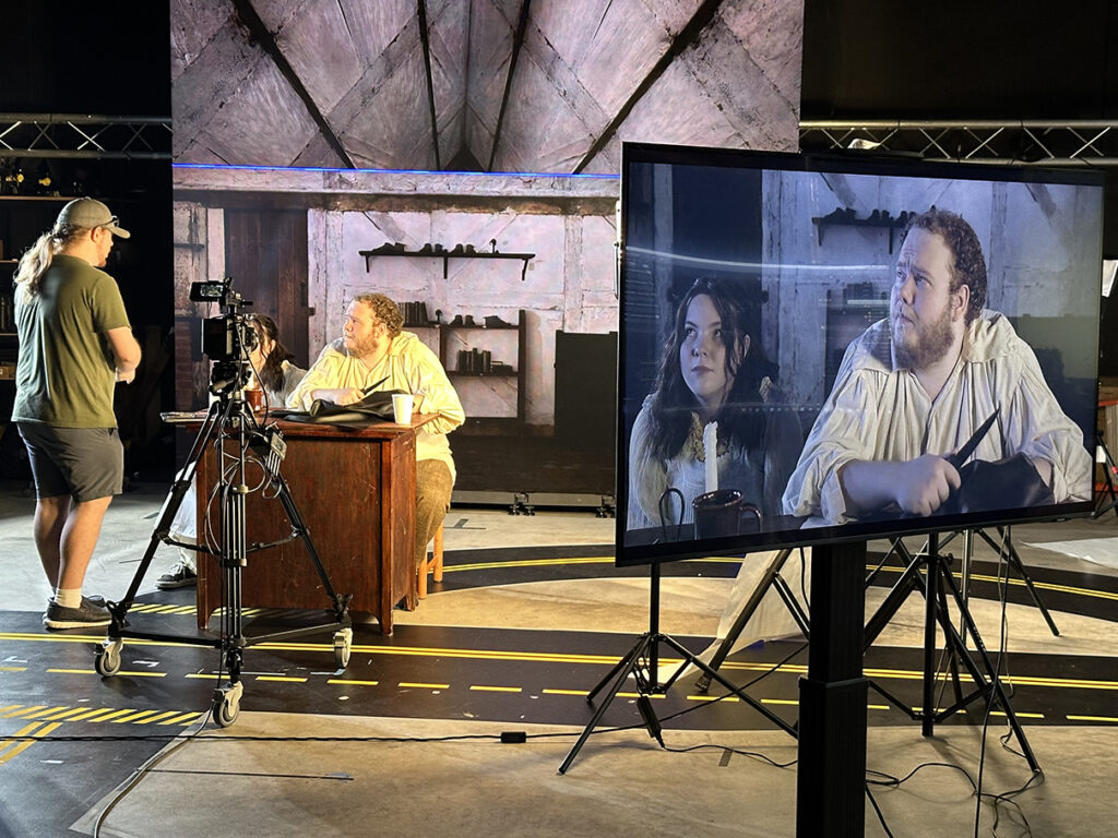 College students work on a set for short film powered by virtual production technology. A large LED screen is in the background, showing the interior of a house. A TV screen at the front right shows the actors in front of the background.