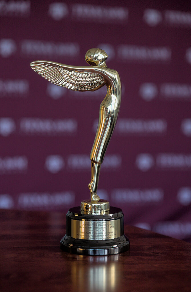 A gold statue in the form of a figure with wings is shown on a table. Behind it is a maroon backdrop with Texas A&M University labeled and its logo included.