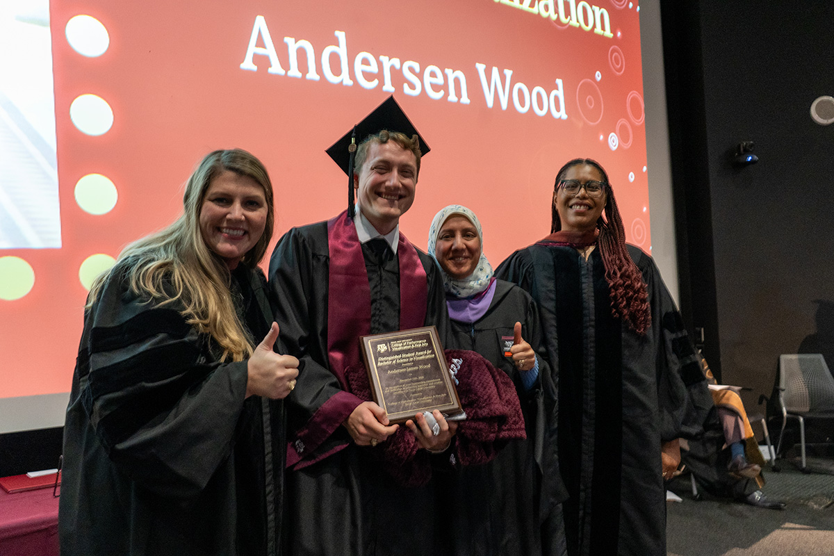 A college student wearing a graduation cap and gown holds a plaque, while three professors wearing graduation gowns stand beside him.