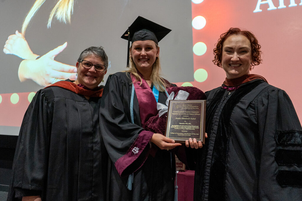A college student in a graduation cap and gown holds a plaque, while two professors wearing caps and gowns are on either side of her.