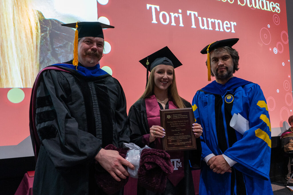 A college student in a graduation cap and gown holds a plaque, while two professors wearing caps and gowns are on either side of her.
