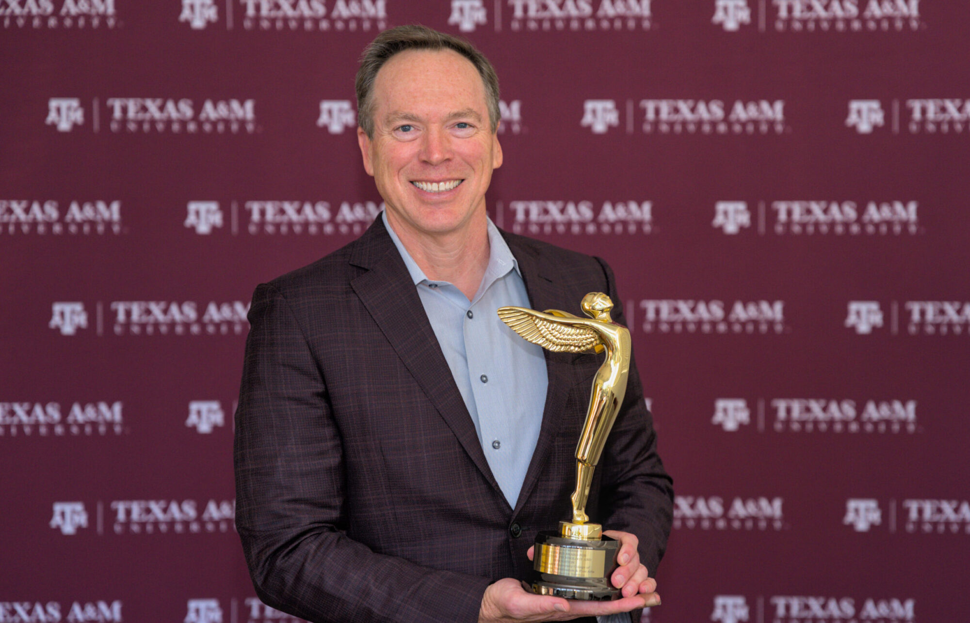 A university dean smiles as he holds a gold trophy in the shape of a figure with wings. Behind him is a maroon backdrop with Texas A&M University and its logo included.