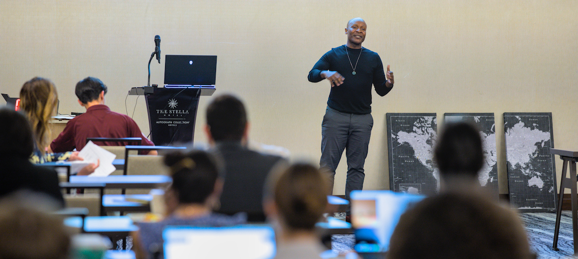 A man stands by a podium, giving an presentation to a group of university faculty members.